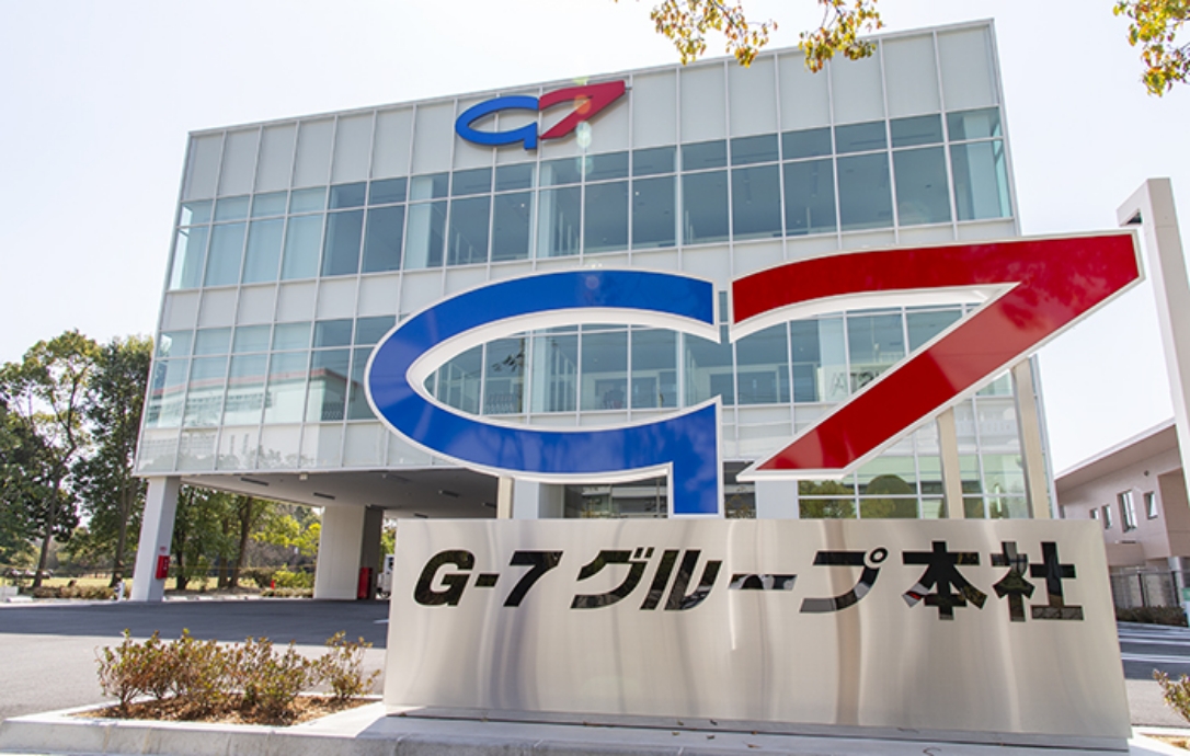 G-7 グループ本社の外観の写真です。G-7 グループのロゴマークの大きい看板が手前に写っています。