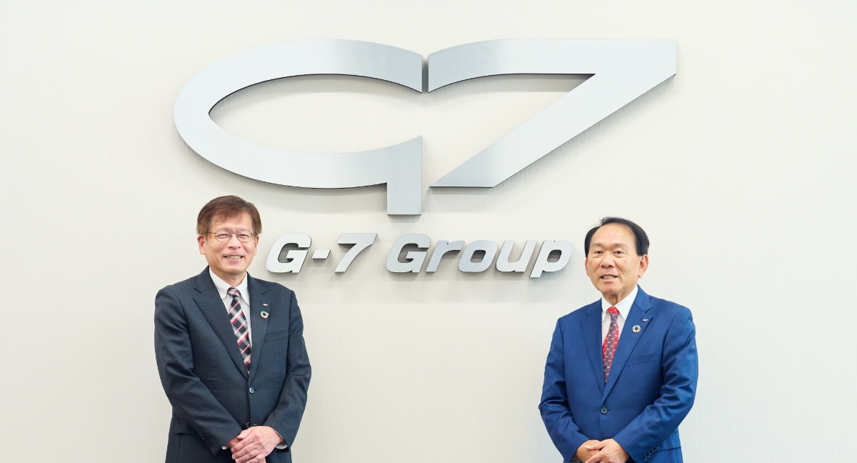 社内の壁面に大きなG-7グループのロゴがあり、その前に株式会社G-7ホールディングス 代表取締役会長兼CEO　金田　達三と代表取締役社長　岸本　安正が並んでいます。