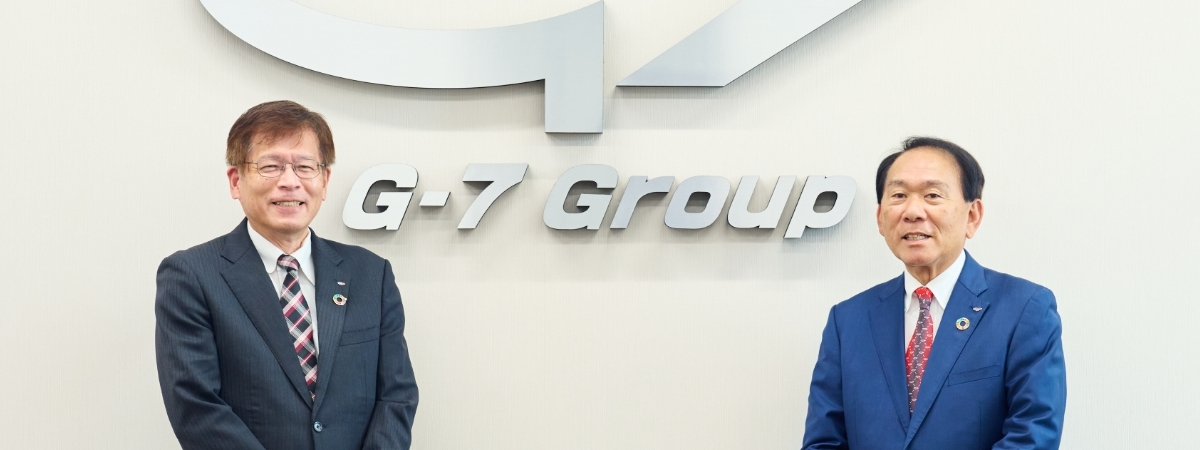 社内の壁面に大きなG-7グループのロゴがあり、その前に株式会社G-7ホールディングス 代表取締役会長兼CEO 金田 達三と代表取締役社長 岸本 安正が並んでいます。